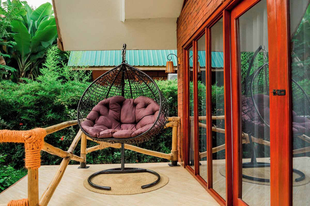 Una silla colgante con asiento acolchado y estructura de metal, ubicada sobre una terraza de madera con un tapete tejido debajo.