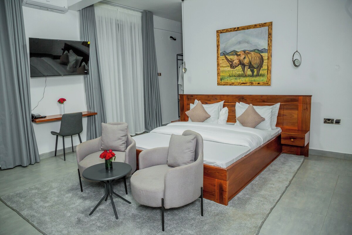 A hotel room with a large bed, two armchairs, a small table with a vase, a TV, and a painting of a rhinoceros.