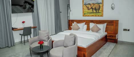 Habitación de hotel con una cama grande, dos sillones, una mesita con un jarrón, un televisor y un cuadro de un rinoceronte.