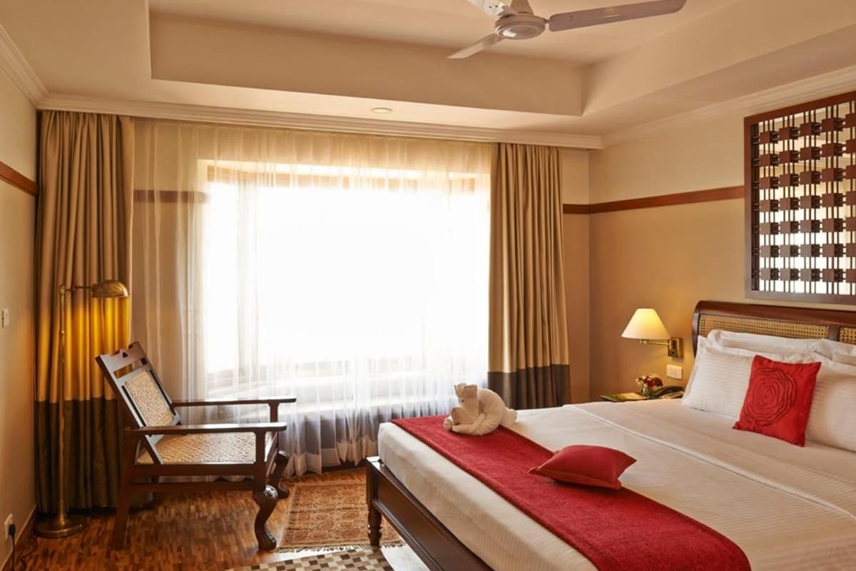 Habitación de hotel con cama, una silla, escritorio, lámpara, ventilador de techo y ventana con cortinas.