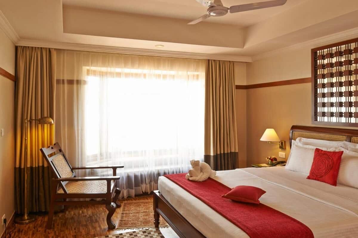 Habitación de hotel con cama, una silla, escritorio, lámpara, ventilador de techo y ventana con cortinas.