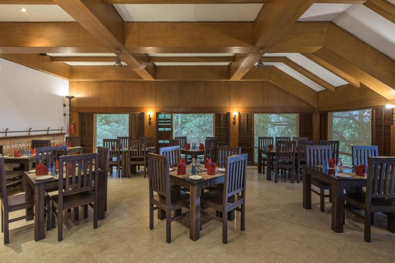 A spacious dining area with wooden tables and chairs, large windows with blinds, and a high ceiling with wooden beams.