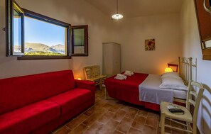Ein Zimmer mit rotem Sofa, zwei Einzelbetten, einem kleinen Tisch, einem Sessel, einem Kleiderschrank und einem Fenster mit Blick auf die Berge.