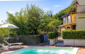 Ein Schwimmbecken mit Sonnenschirm, Liegestuhl und Gartenanlage mit Bäumen und einem Gebäude im Hintergrund.