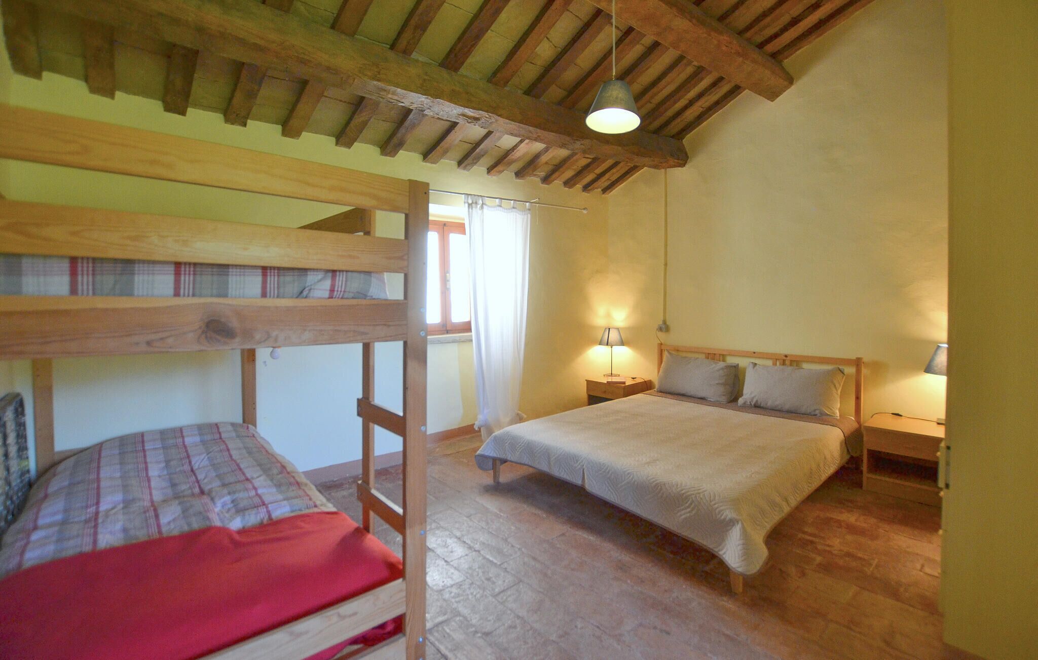 A room with two bunk beds, a single bed, wooden beams on the ceiling, a window with curtains, and a nightstand with a lamp.