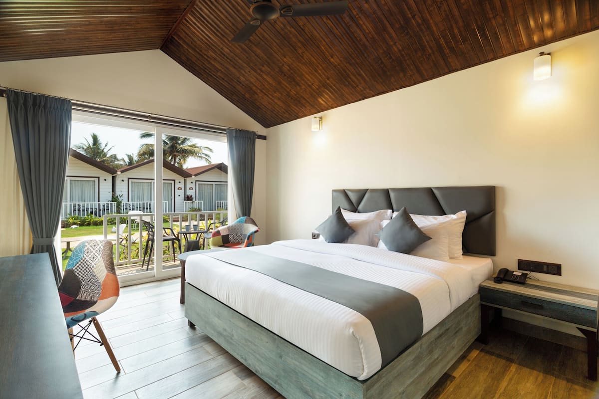 A hotel room with a large bed, a desk, and a chair. There is a balcony with a view of palm trees and a pool.
