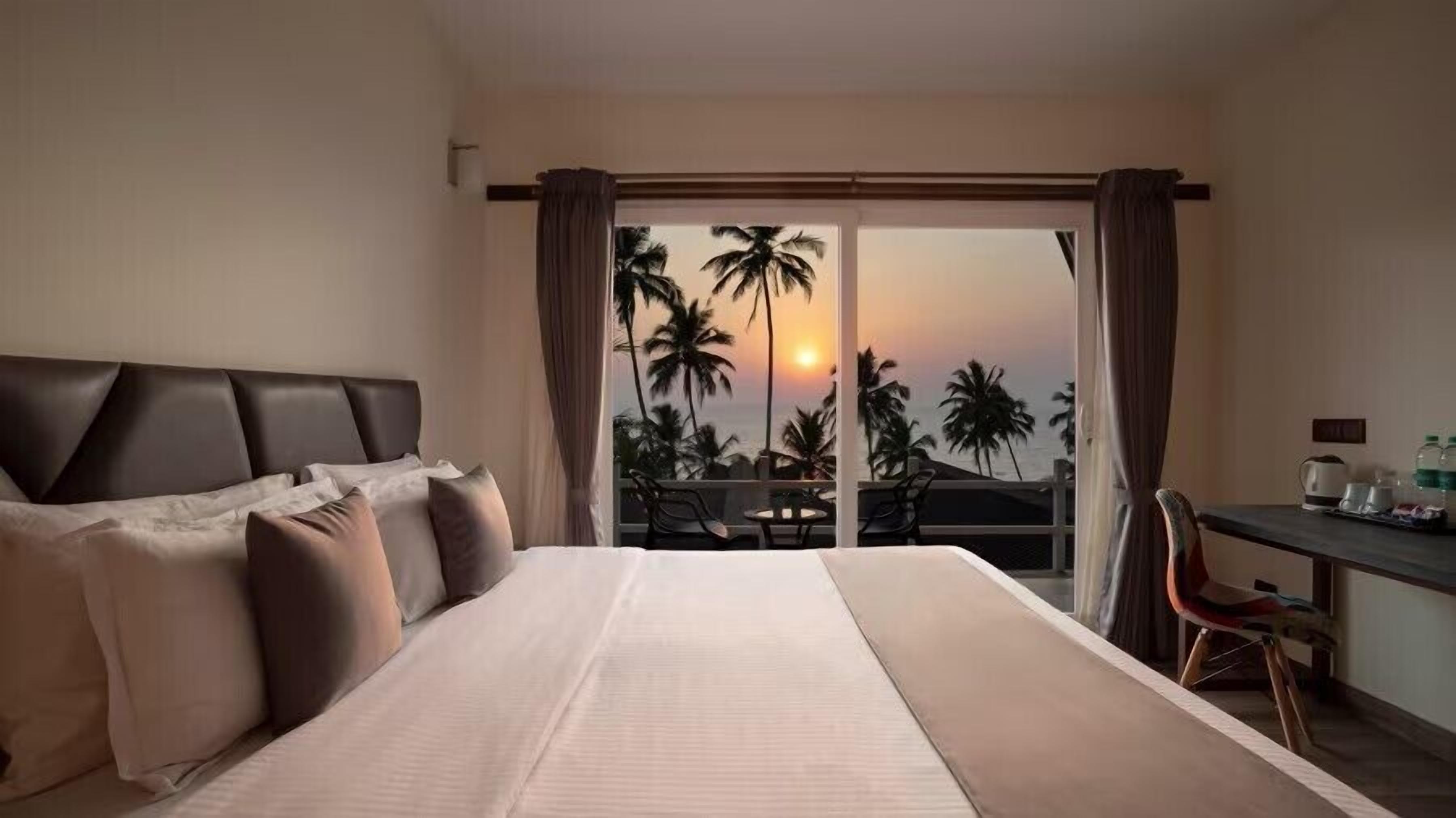A hotel room with a bed, a desk, and a view of palm trees and the sunset.