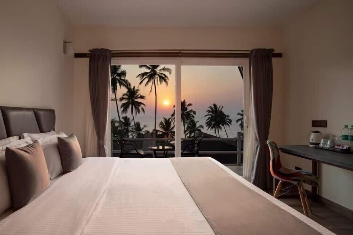 A hotel room with a bed, a desk, and a view of palm trees and the sunset.