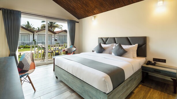 A hotel room with a large bed, a desk, and a chair. There is a balcony with a view of palm trees and a pool. - Stone Wood Beach Resort- A Party Hotel (Vagator)