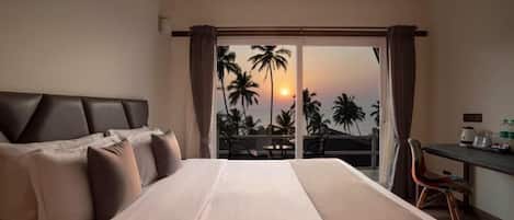 A hotel room with a bed, a desk, and a view of palm trees and the sunset.
