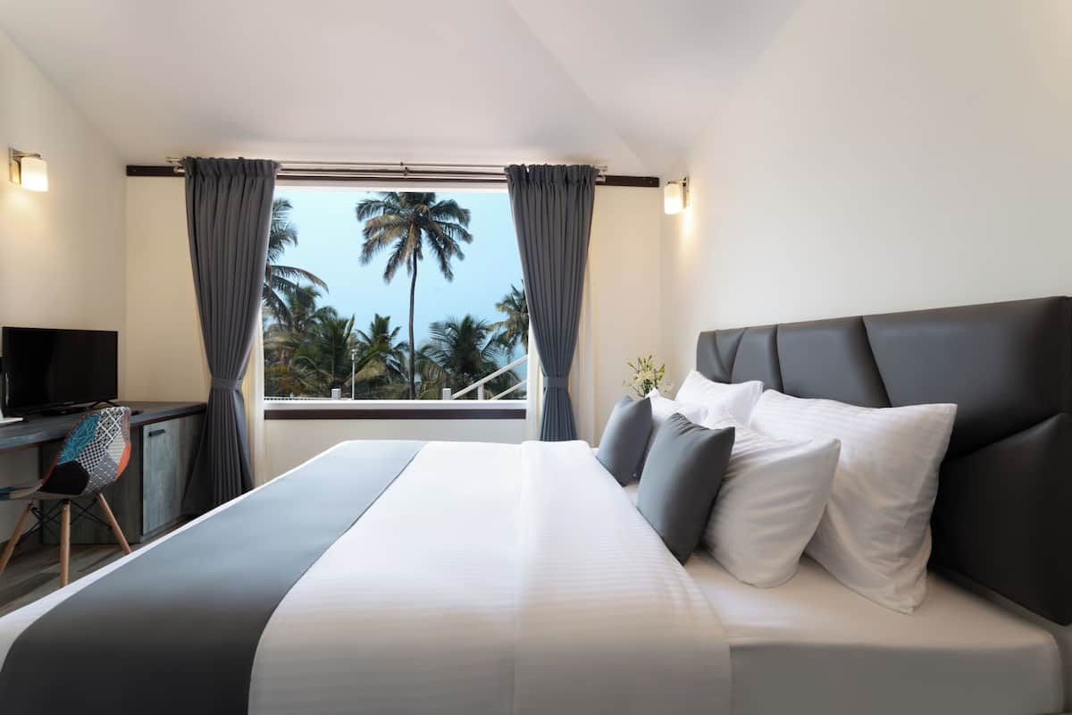 A modern hotel room with a large bed, a desk with a television, and a view of palm trees through the window.