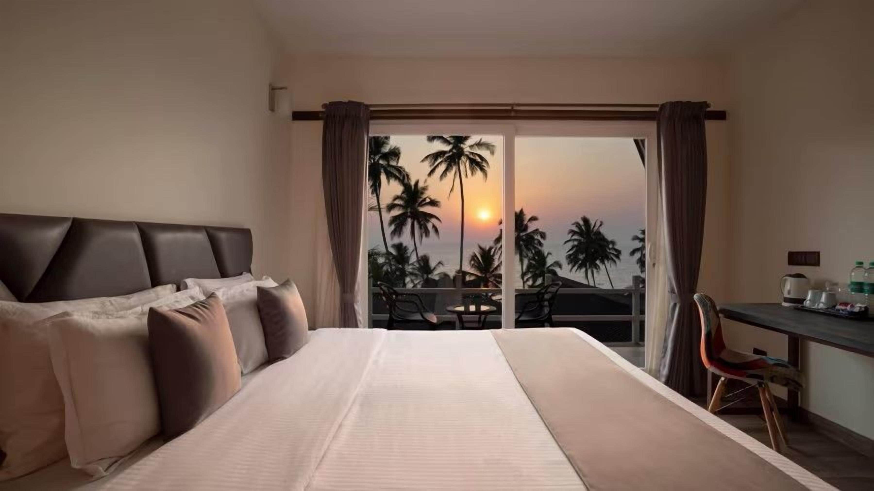 A hotel room with a bed, a desk, and a view of palm trees and the sunset.