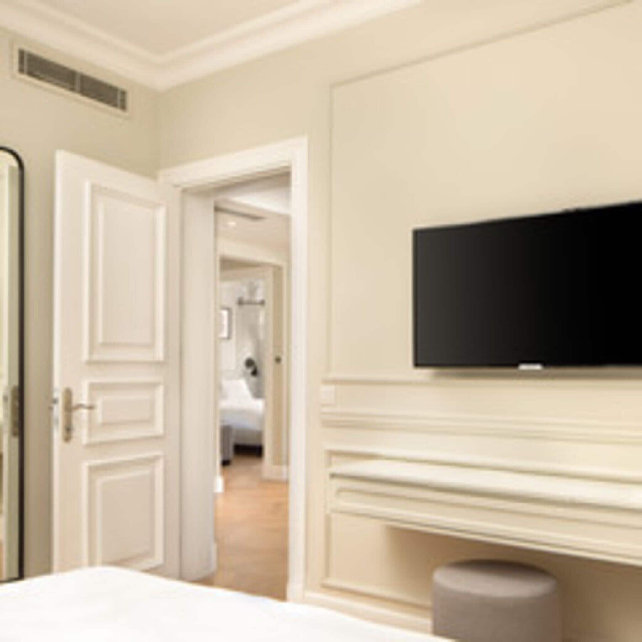 Hotellihuone, jossa on sänky, seinälle asennettu televisio ja ovi, joka johtaa toiseen huoneeseen.