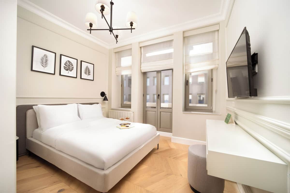 Un dormitorio con cama, mesita de noche, lámpara, un cuadro con hojas y ventanas, además de un televisor.