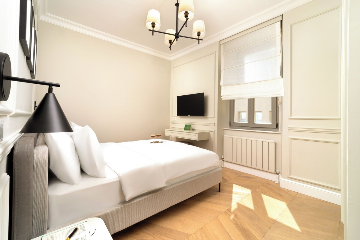 A hotel room with a bed, a TV, a window with blinds, a radiator, and a chandelier.