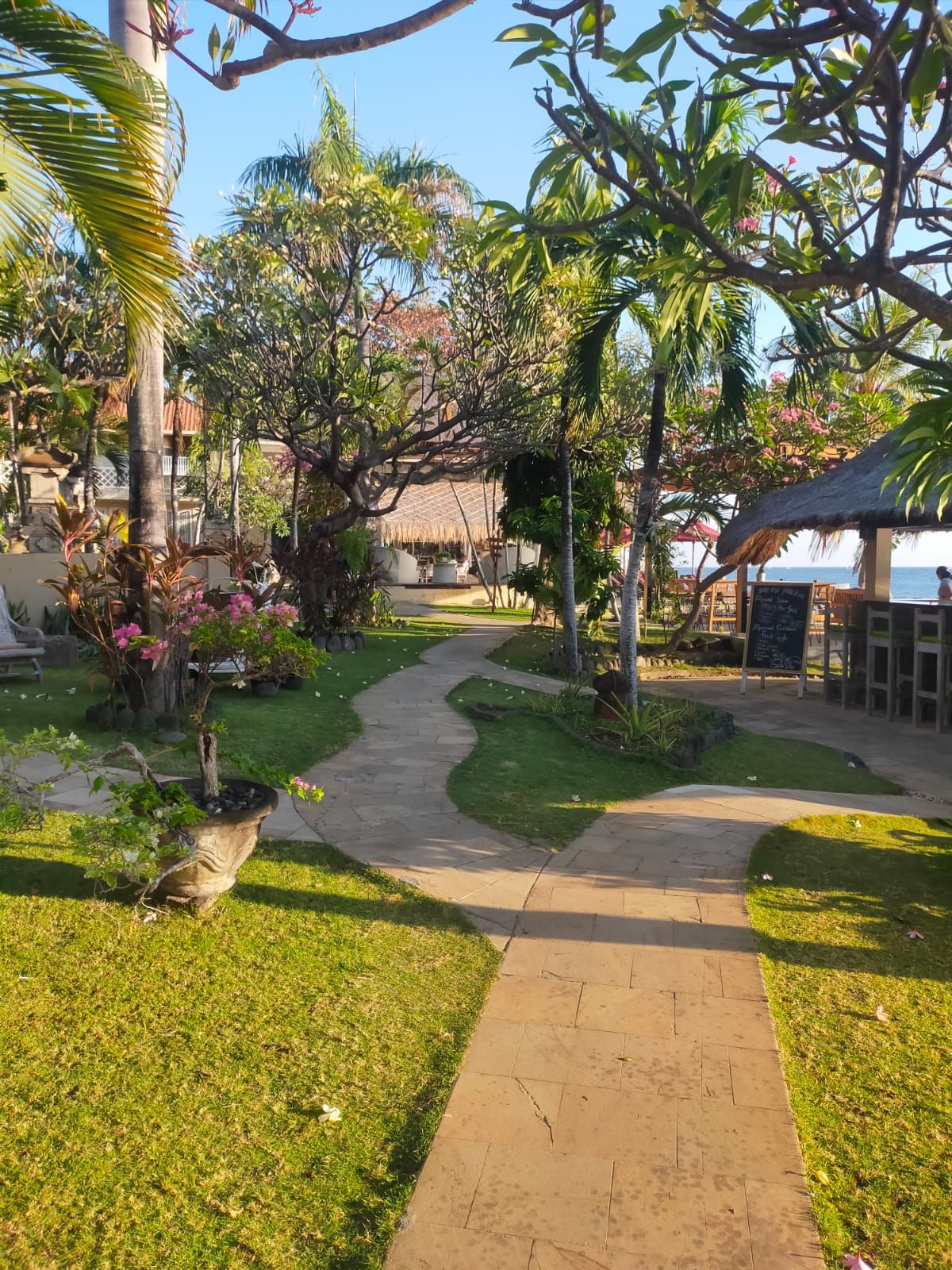 Un sendero de jardín que conduce a un edificio con tejado de paja, rodeado de plantas tropicales y un cielo despejado.