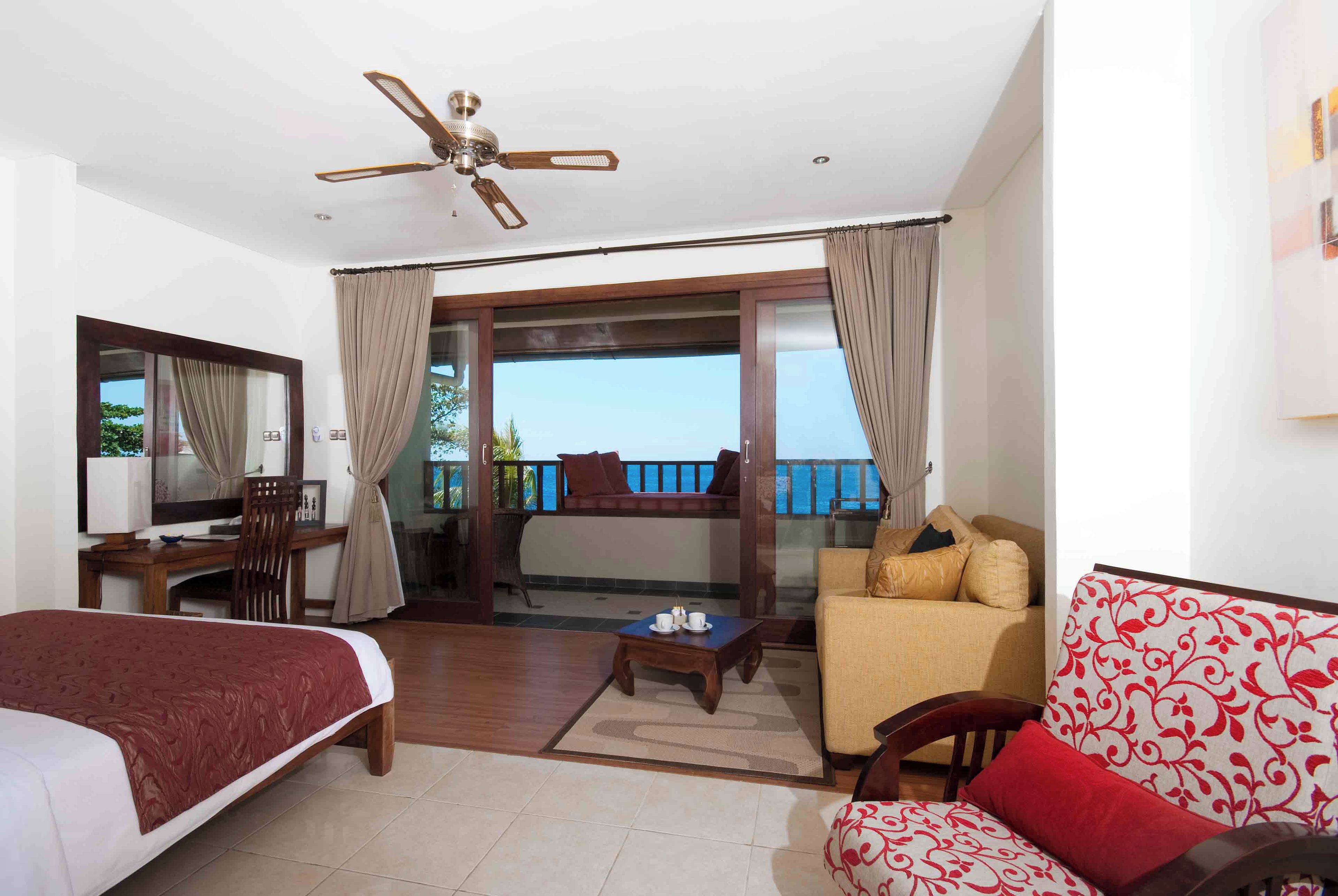 Habitación de hotel con cama, sofá, escritorio, silla, balcón con vistas y ventilador de techo.