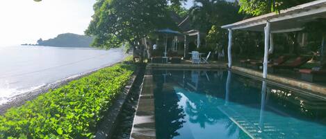 A swimming pool with a view of the sea, surrounded by greenery and a covered seating area.