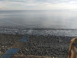 A rocky beach with a rope extending into the sea, a metal grid walkway, and a wooden structure on the right. - Tauch Terminal Resort Tulamben & Spa (Rendang)