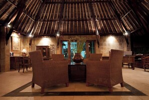Una habitación con techo alto e inclinado, muebles de mimbre, paredes de piedra y una mesa central.