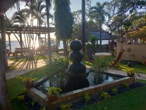 Un jardín con fuente de agua, palmeras y un espacio de descanso junto al agua.