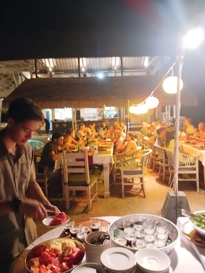 A dining area with tables and chairs, a person serving food, and various dishes and drinks.