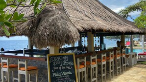 A beachside bar with a thatched roof, wooden bar stools, and a chalkboard menu.