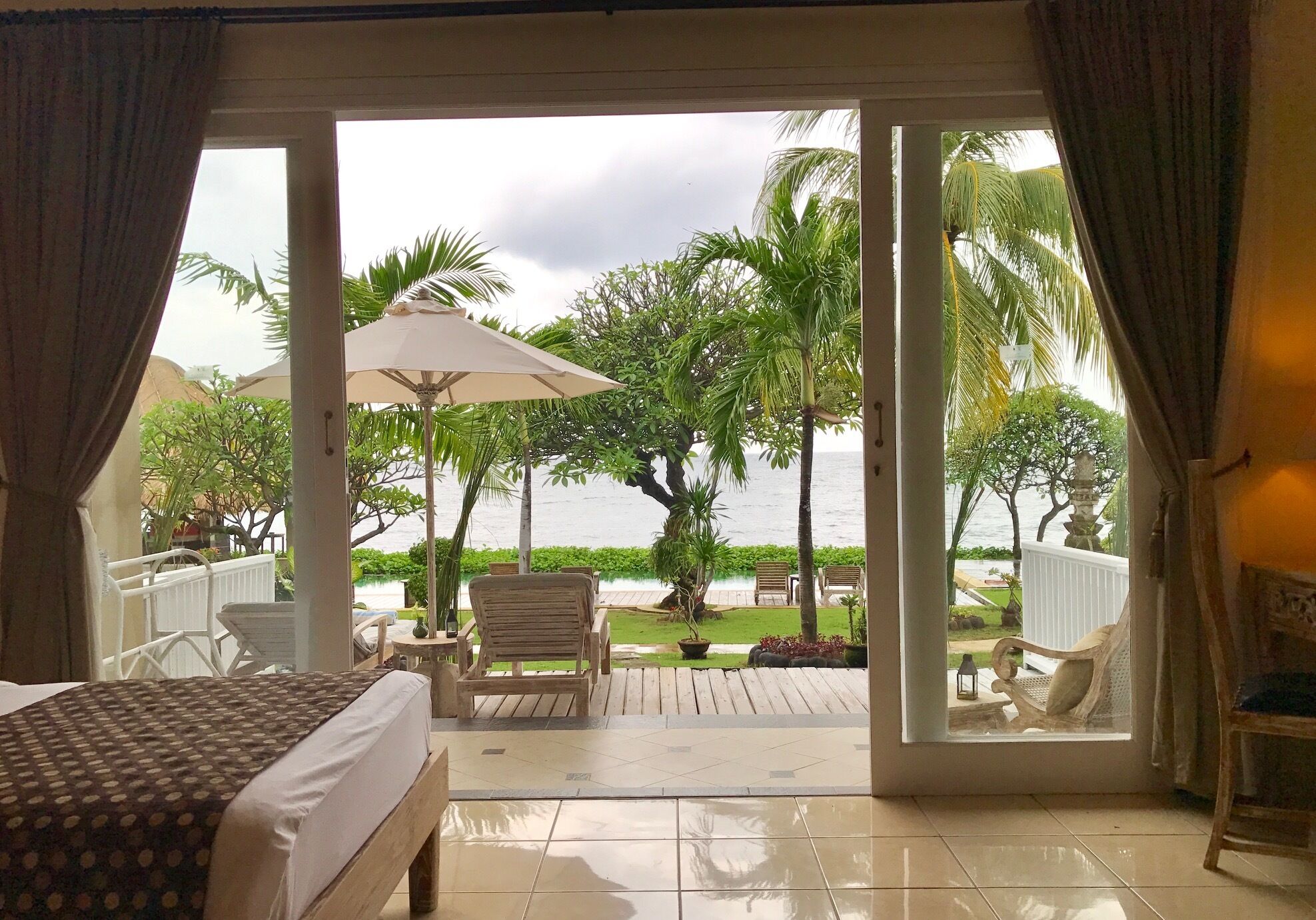 A room with a bed, a view of the outdoors, a patio area with chairs, and a large umbrella.