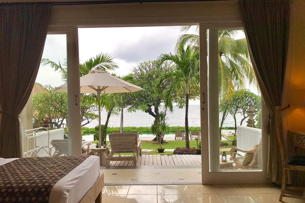 A hotel room with a bed, a wooden chair, a small table, and a view of a pool and palm trees.