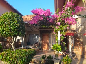 Una propiedad residencial con un porche cubierto, una puerta de madera y un pequeño jardín con diversas plantas.