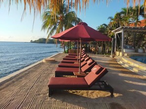 Un espace de détente aménagé au bord de la plage, avec des chaises longues et une piscine, entouré de palmiers et offrant une vue sur l’océan.