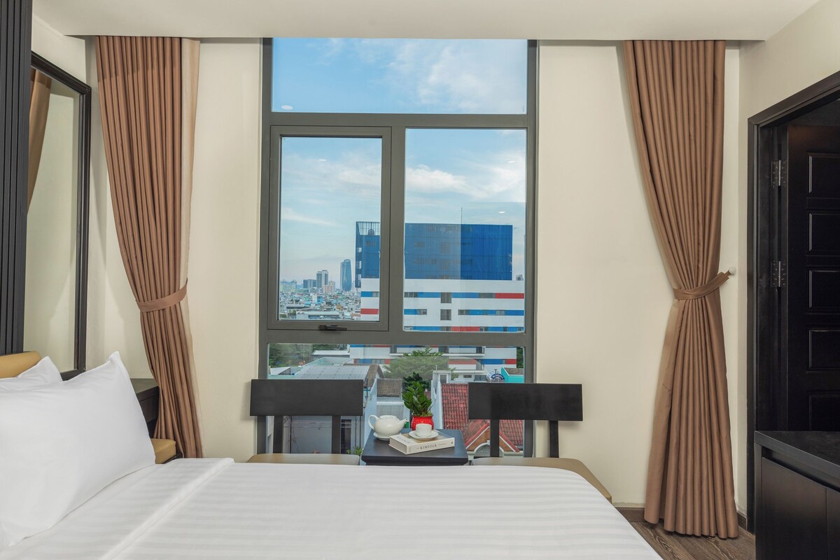 A hotel room with a large window, a bed with white linens, a small table with a teapot and a book, and a view of a cityscape.