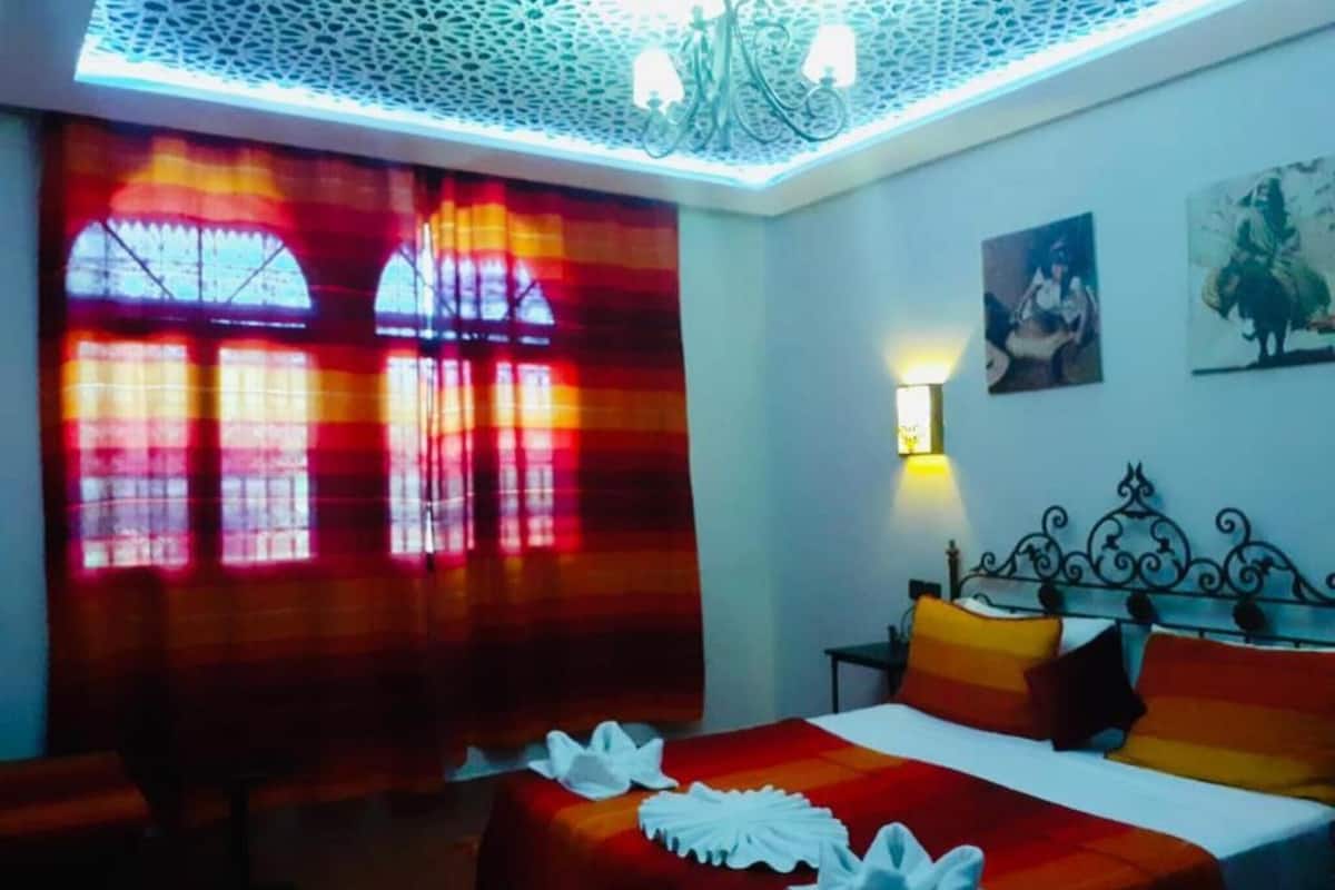 A hotel room with a bed, a bench, a chandelier, and a window with curtains.