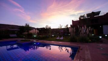 A swimming pool with a tiled deck, surrounded by vegetation and a building with a flat roof.