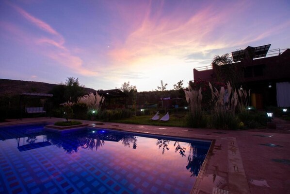 A swimming pool with a tiled deck, surrounded by vegetation and a building with a flat roof.