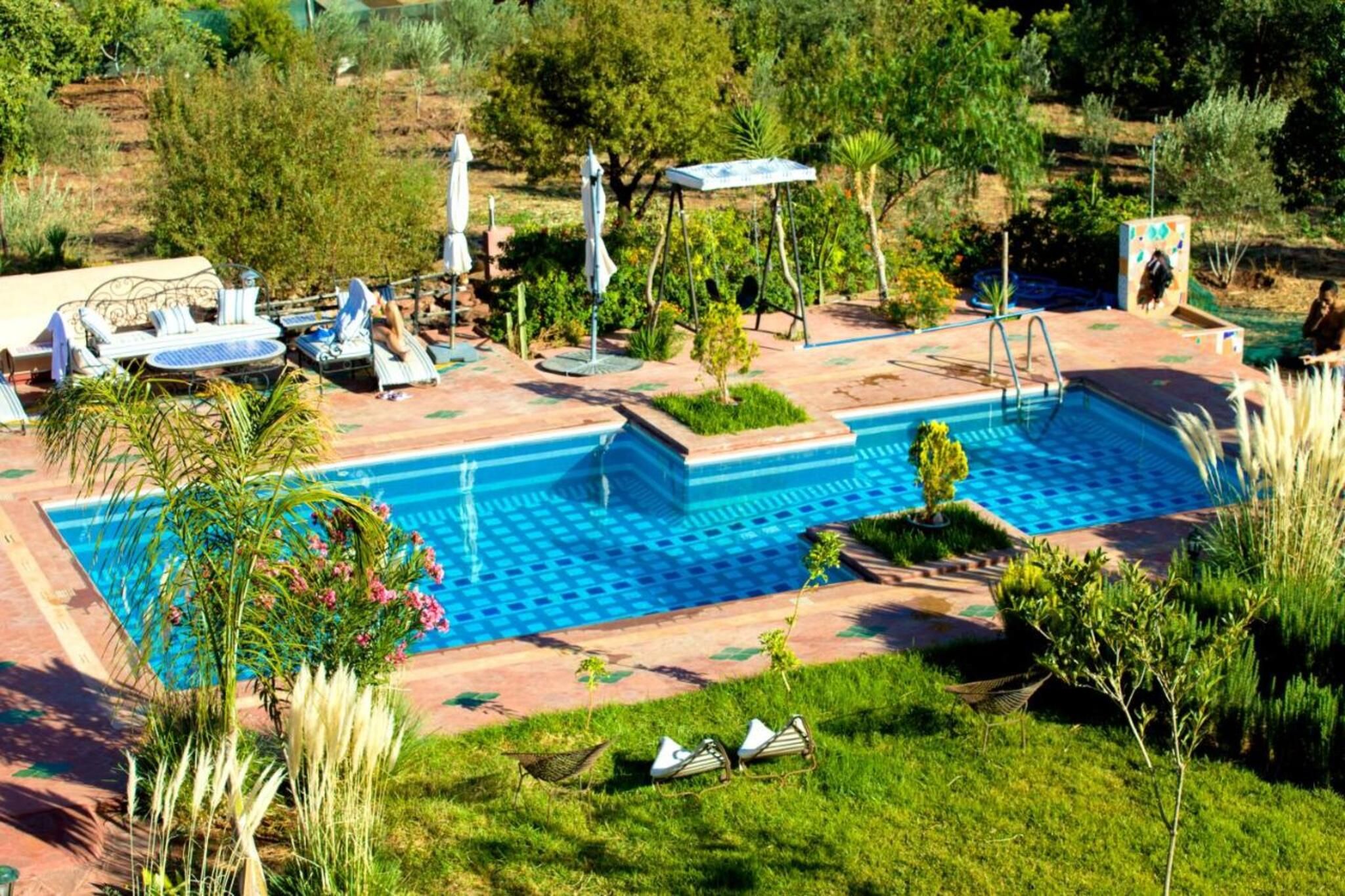 Une piscine avec des chaises longues, un espace de détente couvert et un jardin agrémenté de différentes plantes.