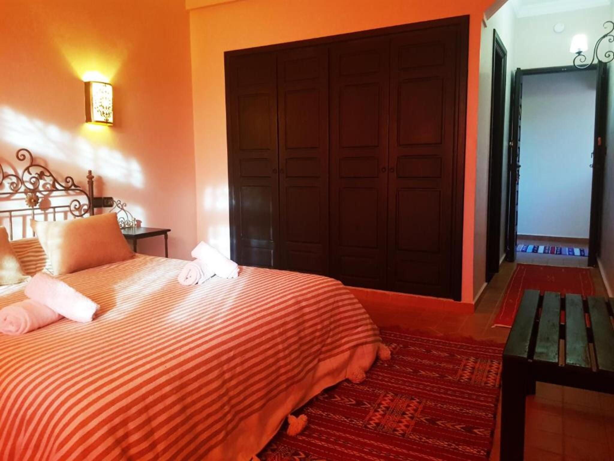 Une chambre à coucher comprenant un lit, une table de chevet, une lampe fixée au mur, une armoire et un banc.