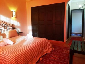 A bedroom with a bed, a nightstand, a wall-mounted light, a wardrobe, and a bench. - Les Jardins Yasmina (Sidi Dahmane)
