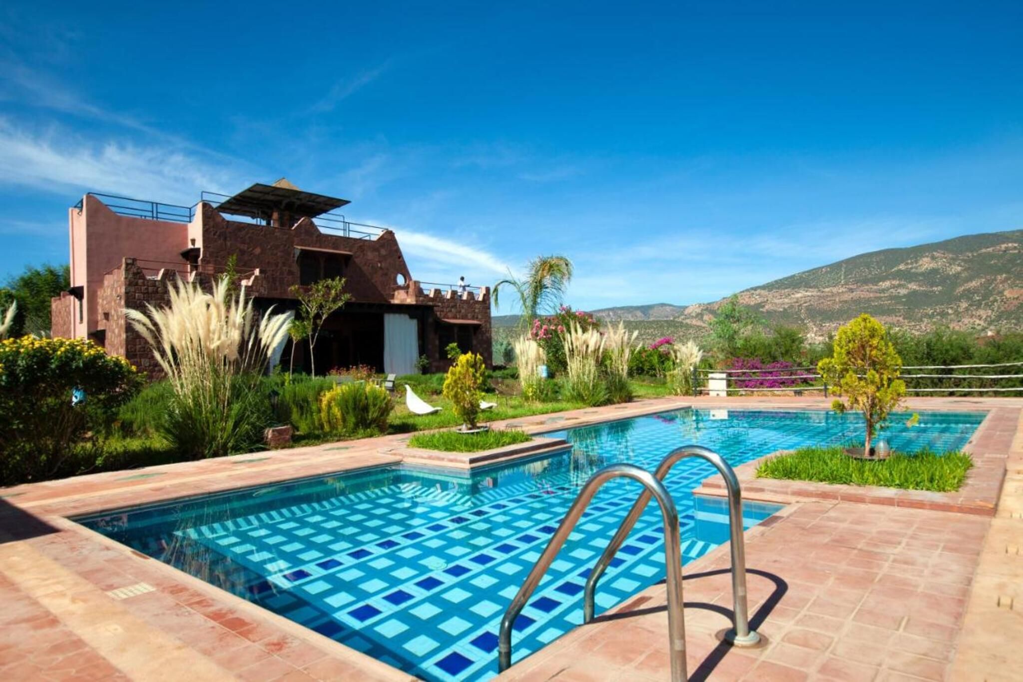 A swimming pool with a geometric tile pattern, surrounded by a landscaped garden with various plants and a view of a mountainous landscape.