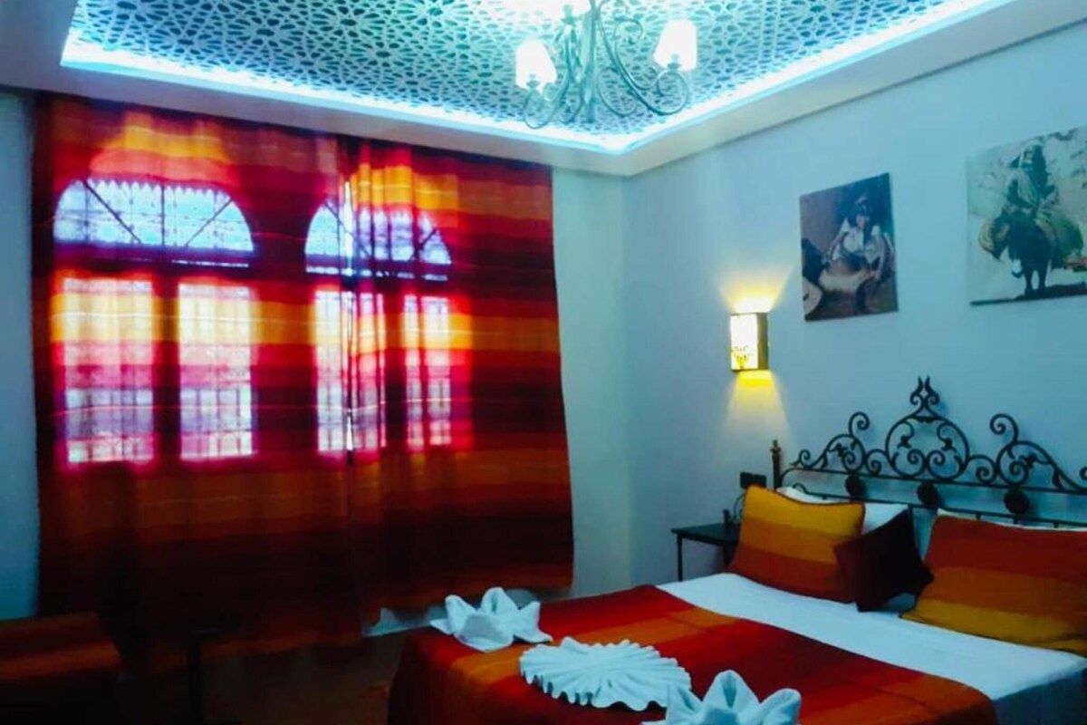 A hotel room with a bed, a bench, a chandelier, and a window with curtains.