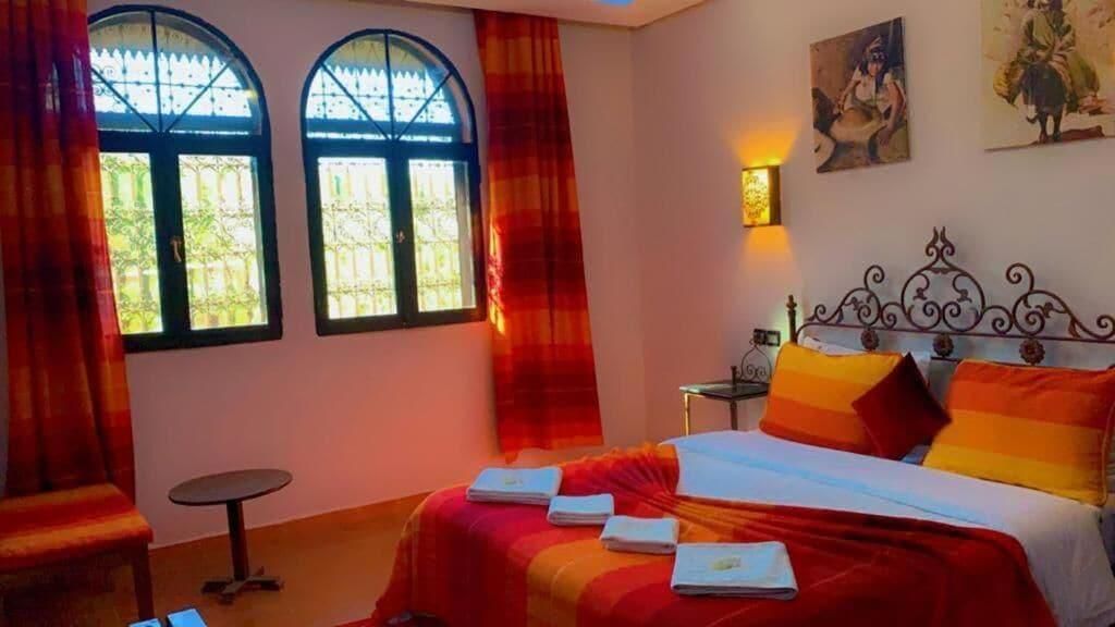 A room with a bed, two windows, a table, and a wall-mounted light.