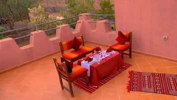 A terrace with a table set for two, two chairs, a red tablecloth, and a patterned rug.