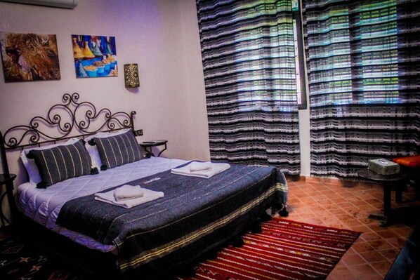 A bed with a metal frame, a red and black striped rug, and a wall with two paintings. - Les Jardins Yasmina (Sidi Dahmane)