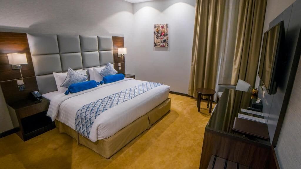 A hotel room with a large bed, bedside tables with lamps, a TV, and a desk with a computer.