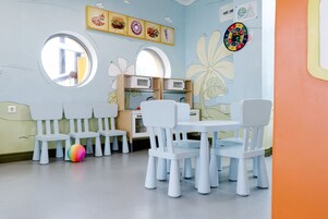 Una sala de juegos infantil con sillas blancas, una pelota de colores y decoraciones en la pared que incluyen ilustraciones de comida y un dardo.