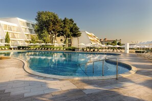 A swimming pool with a curved design, surrounded by lounge chairs and umbrellas, with a modern building in the background. - Aluasun Helios Beach (Nessebar)