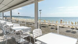 A beachside restaurant with outdoor seating, large windows, and a view of the beach and sea. - Aluasun Helios Beach (Nessebar)