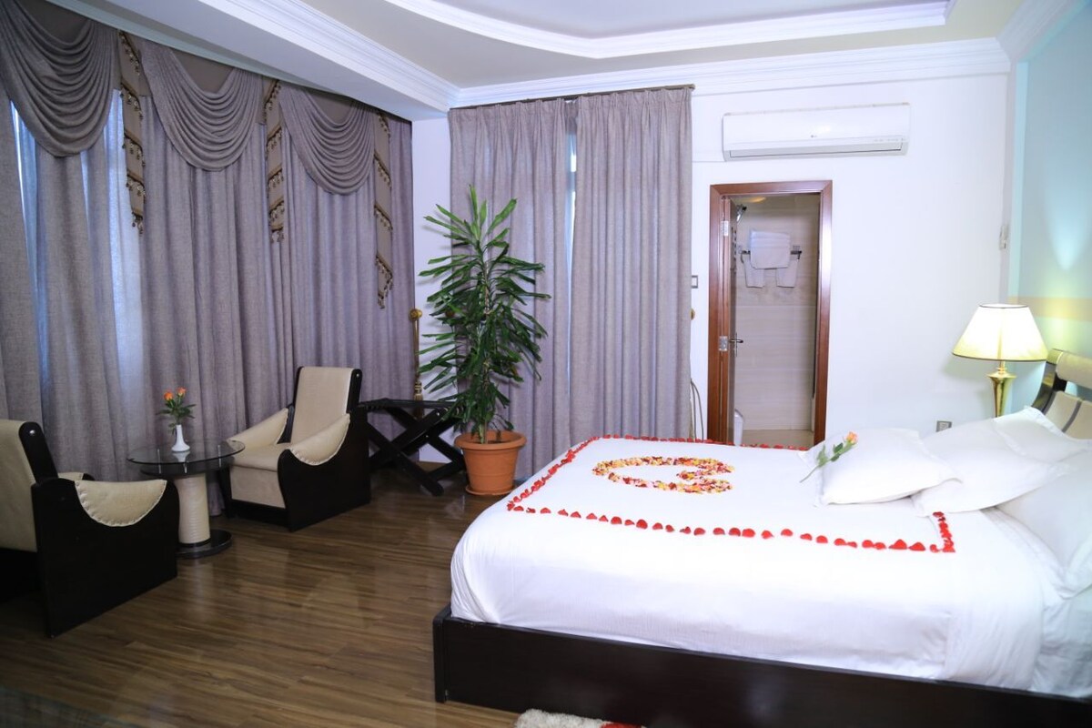 Habitación de hotel con cama, una silla, una mesa, una planta en maceta y una puerta que da a un baño.