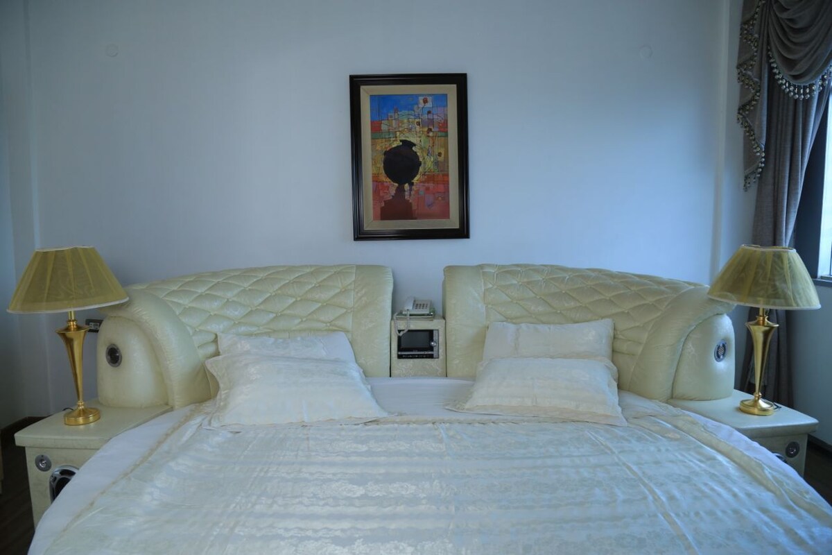 Una cama doble con cabecera acolchada, lámparas de noche, una mesita auxiliar pequeña, una obra de arte enmarcada en la pared y una ventana con cortinas.