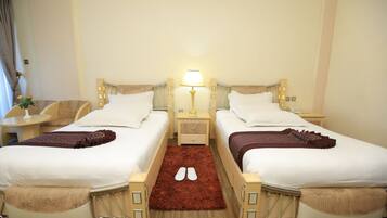 A hotel room with two beds, a nightstand with a lamp, a small table with a vase, and a red rug.